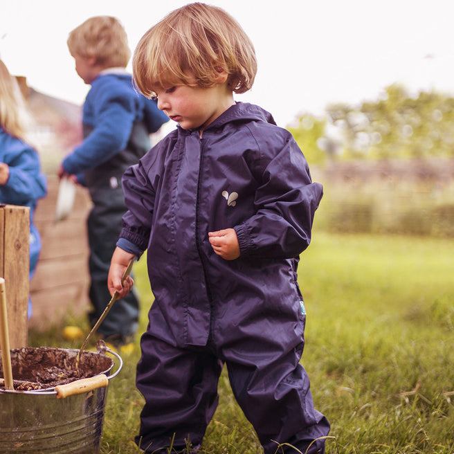 A child wearing the 'Originals Waterproof Puddle Suit Navy' playing with a stick in a bucket outdoors on a grassy field.